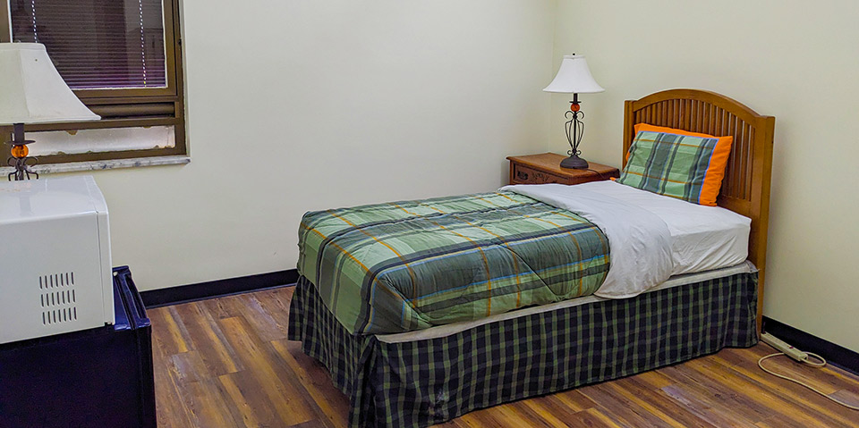 Image of dormitory room on the new Woodlands Campus. Shown is a single bed, with nightstand and lamp.
